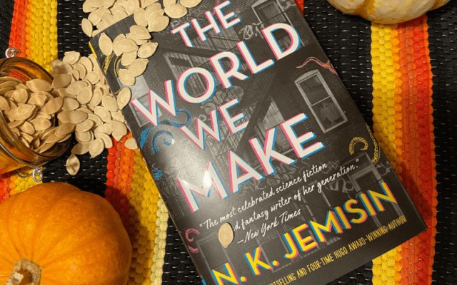 A colorful image of the book "The World We Make" by N.K. Jemisin, surrounded by pumpkins and fall de.