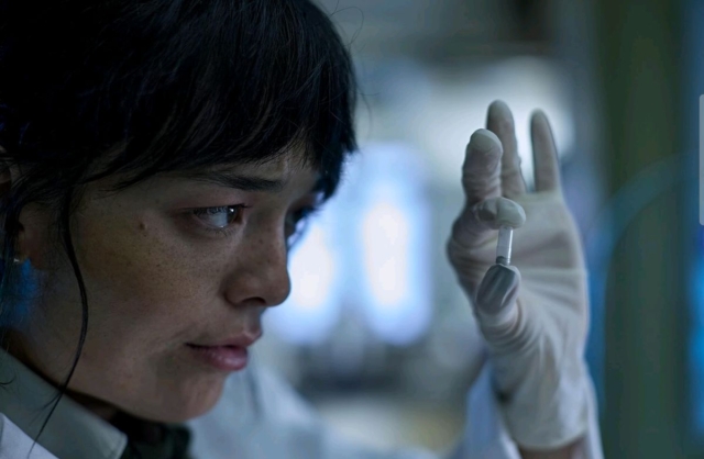 Midori Francis in Saccharine A woman scientist holding a syringe in a laboratory setting, focused on her work.