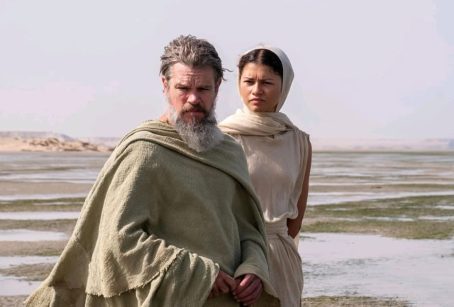 Two people in historical costumes on a beach with tide pools and a cloudy sky. Matt Damon and Zendaya in The Odyssey