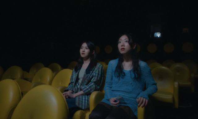 Two women sitting in a dark theater with yellow seats, watching a film. Still from You Are the Film