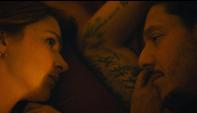 A still from Bruton, a 2026 film. Intimate close-up of a couple gazing at each other in a dimly lit setting.