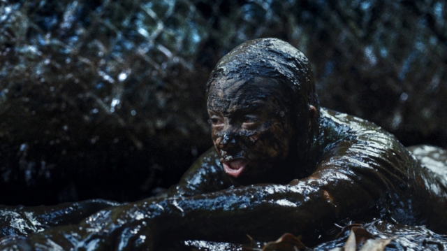 A scene from the 2026 film Beware Boiuna. Child covered in mud crawling through a forest stream.