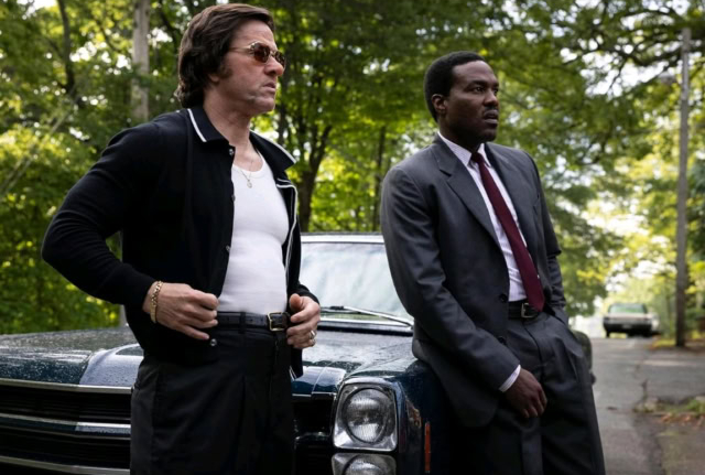 Two men in suits standing outdoors near a black car in a park setting. Mark Wahlberg and Yahya Abdul-Mateen II in the film By Any Means