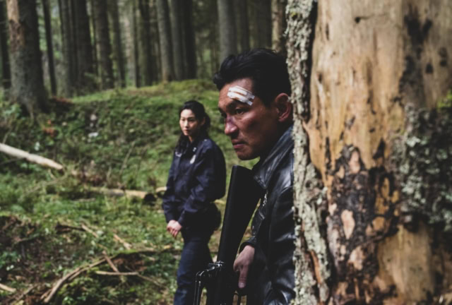 Two people in a forest, one hiding behind a tree, the other standing nearby.