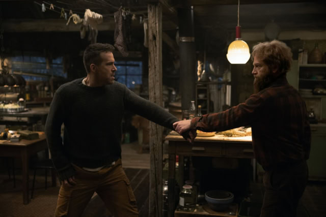 Two men in a tense confrontation in a rustic, dimly lit workshop. Scene from the film Mayday on Apple TV starring Ryan Reynolds and Kenneth Branagh