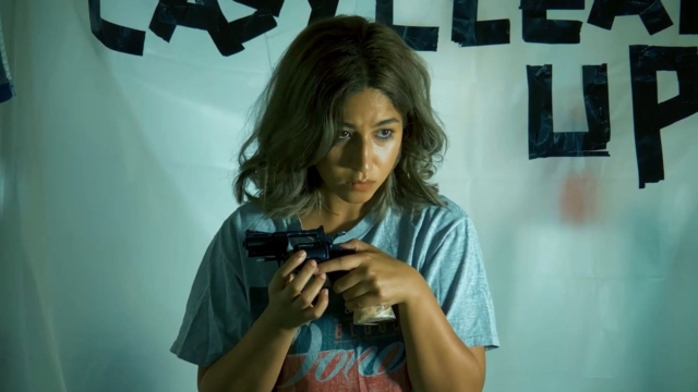 From the film Contemplation by Curry Barker. Woman holding a gun in a room with a banner in the background.