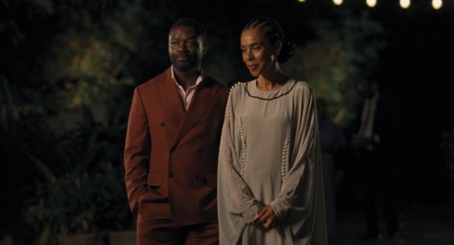 Two people standing outdoors at night, dressed elegantly, with trees in the background.