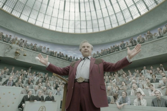 A man in a maroon suit speaking in a large, domed courtroom filled with spectators.