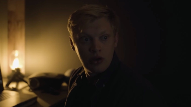 From the film Heavy Eyes by Curry Barker. Young man in a dark room with a surprised expression, illuminated by warm light.